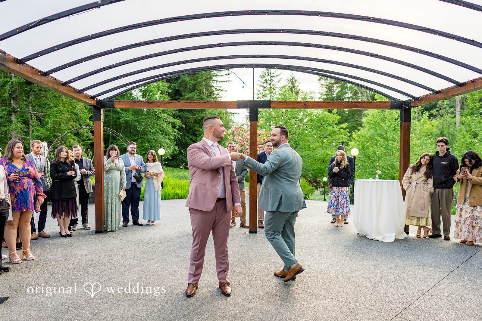 Kristian + Jeff Couple dancing together under a covered outdoor pavilion with wedding guests watching.