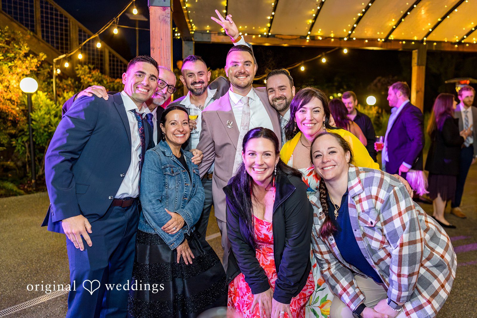 Kristian + Jeff A smiling group of wedding guests posing closely together for a photo at night.