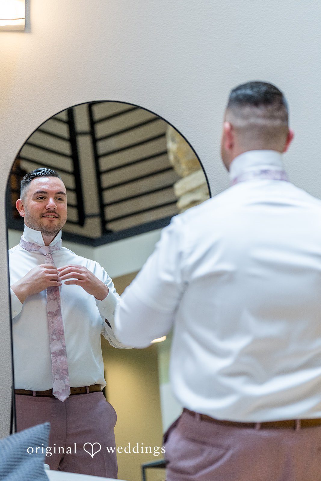 Kristian + Jeff A man in a white shirt adjusting his tie while looking into a large round wall mirror.