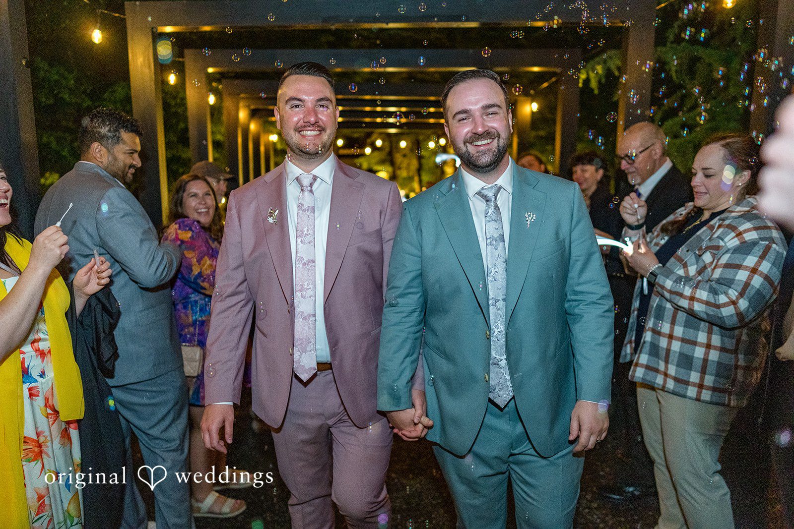 Kristian + Jeff Couple holding hands, smiling, and walking forward through a crowd of applauding guests at night.