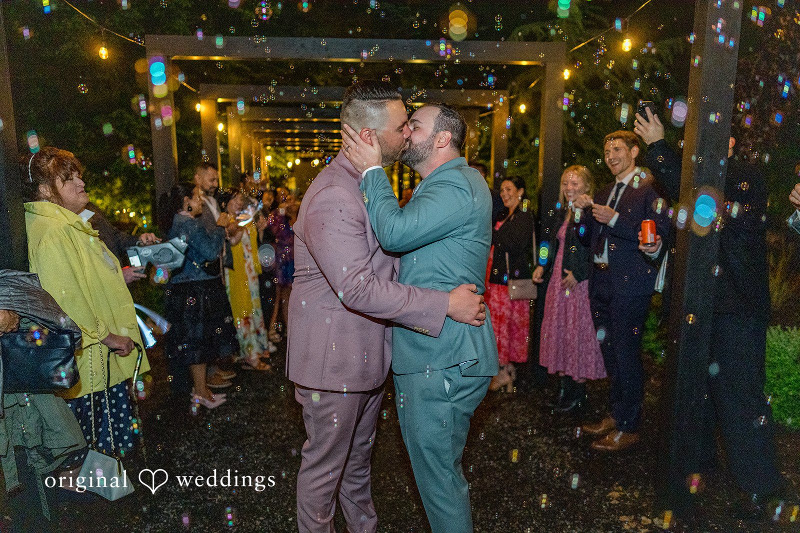 Kristian + Jeff Couple sharing a kiss at night while guests surround them.