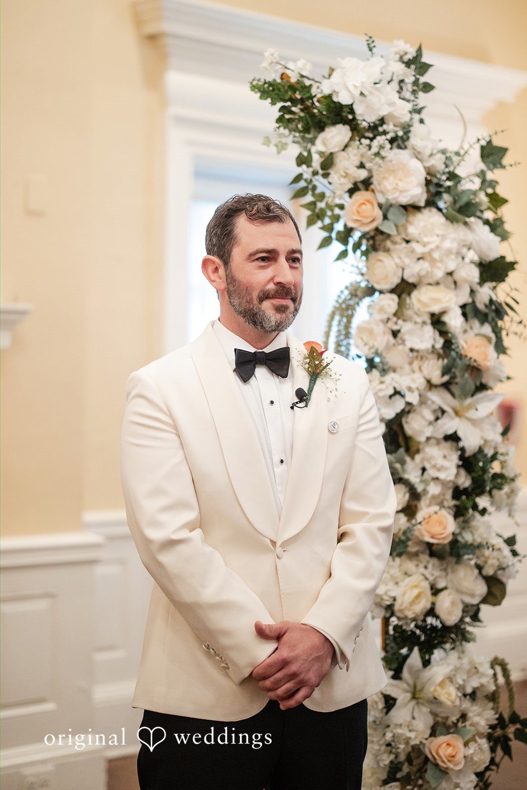Groom smiling while standing in front of a white floral wedding arch.