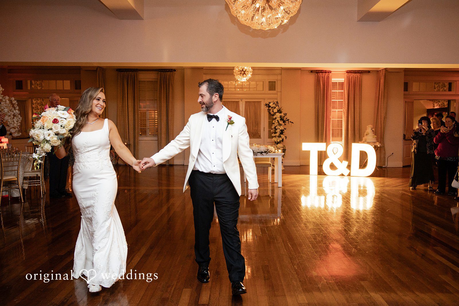 Bride and groom holding hands on a dance floor illuminated by large "T&D" marquee letters.