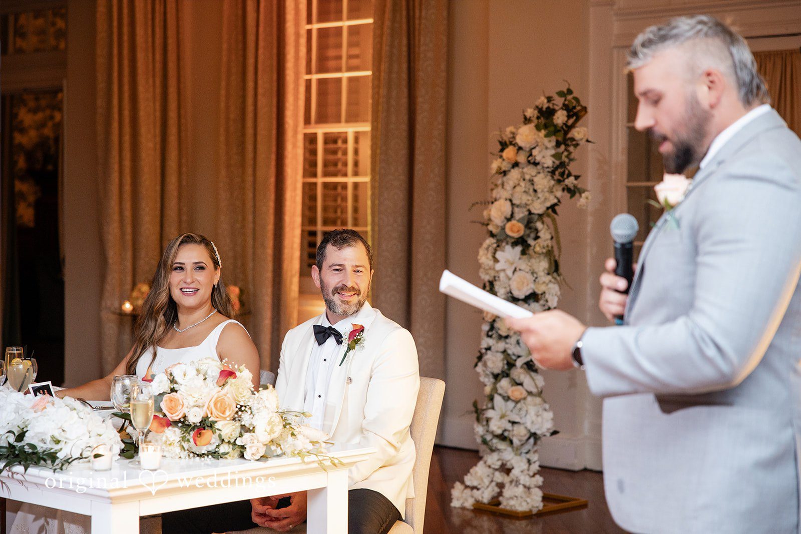 Man holding a paper and speaking into a microphone to toast the seated bride and groom.