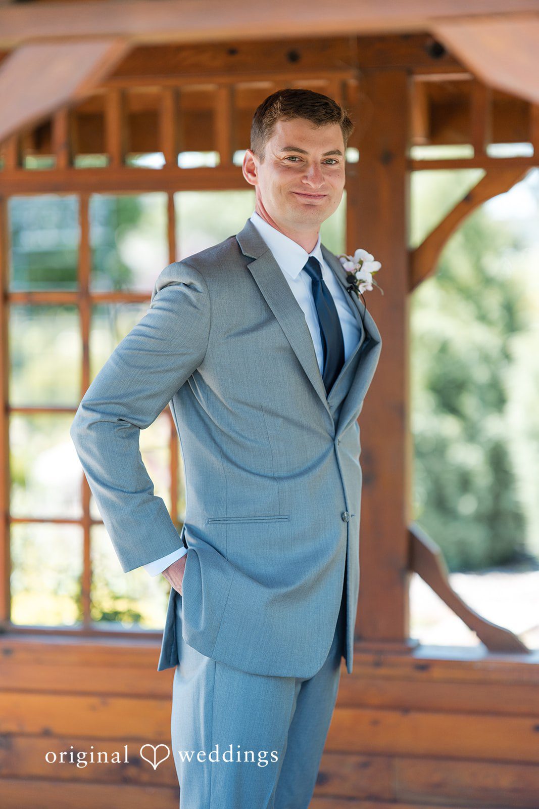 Cassandra + Adam Groom in a light grey three-piece suit posing by a wooden gazebo.