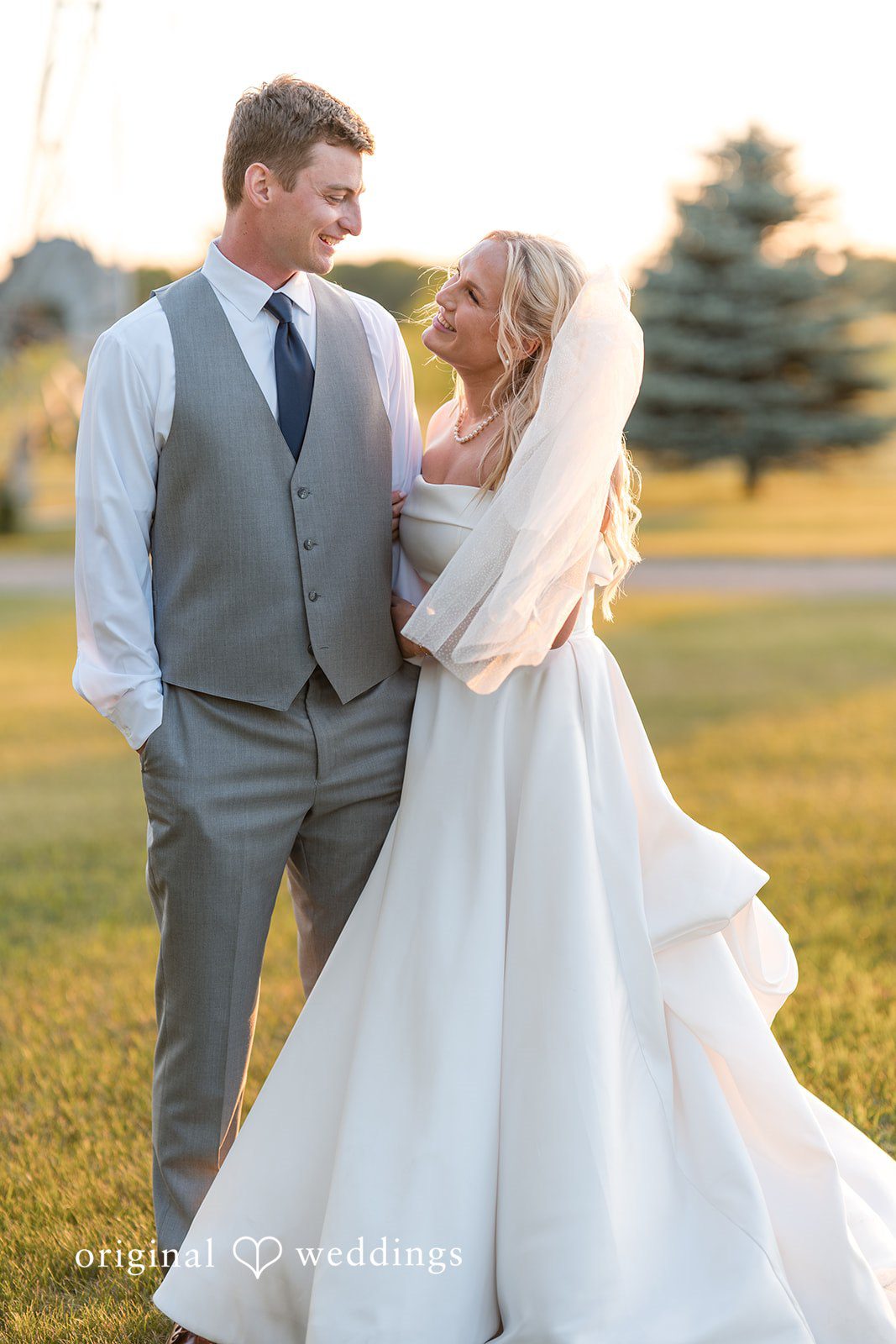 Cassandra + Adam Groom wrapping his arms around the bride from behind in a grassy area.
