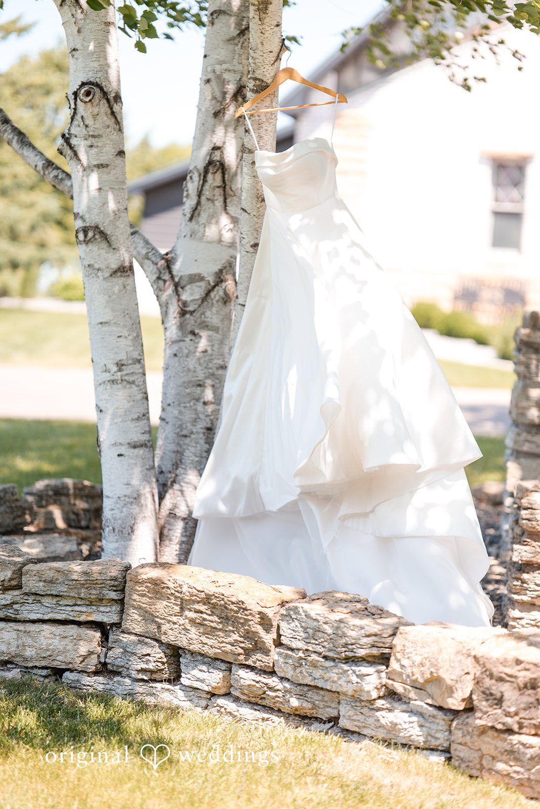 Cassandra + Adam White wedding dress hanging from a tree branch next to a stone wall.