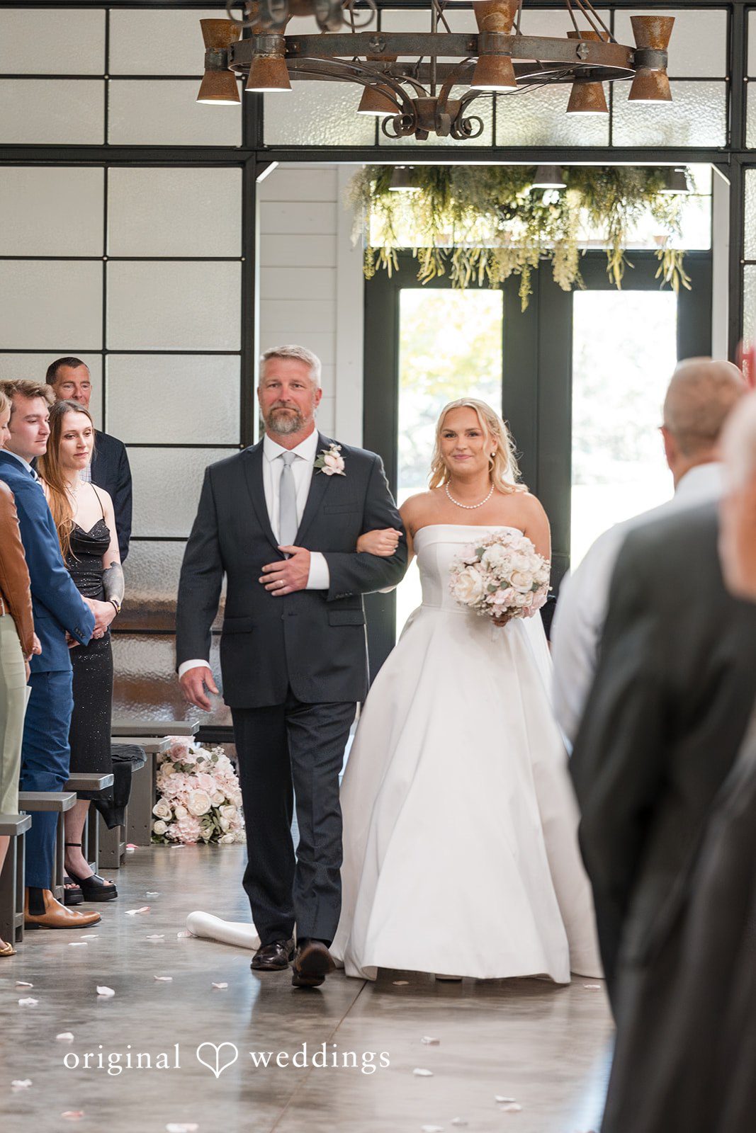 Cassandra + Adam Bride walking down the aisle on a man's arm past seated guests.