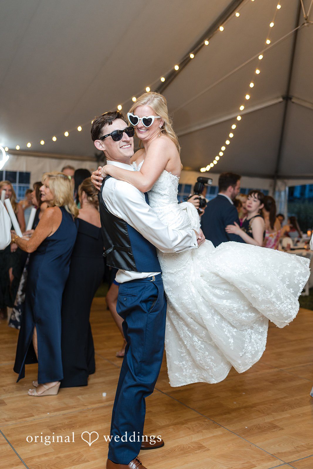 John + Emma Groom lifting the bride into the air while both wear sunglasses on the dance floor.