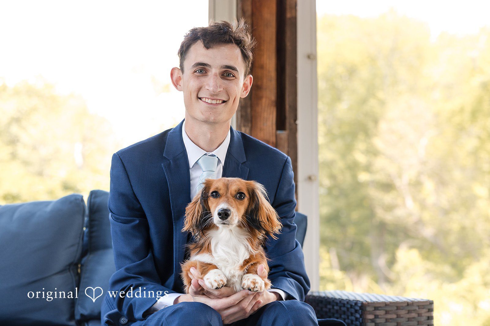 John + Emma Smiling groom sitting down and holding a small brown dog.