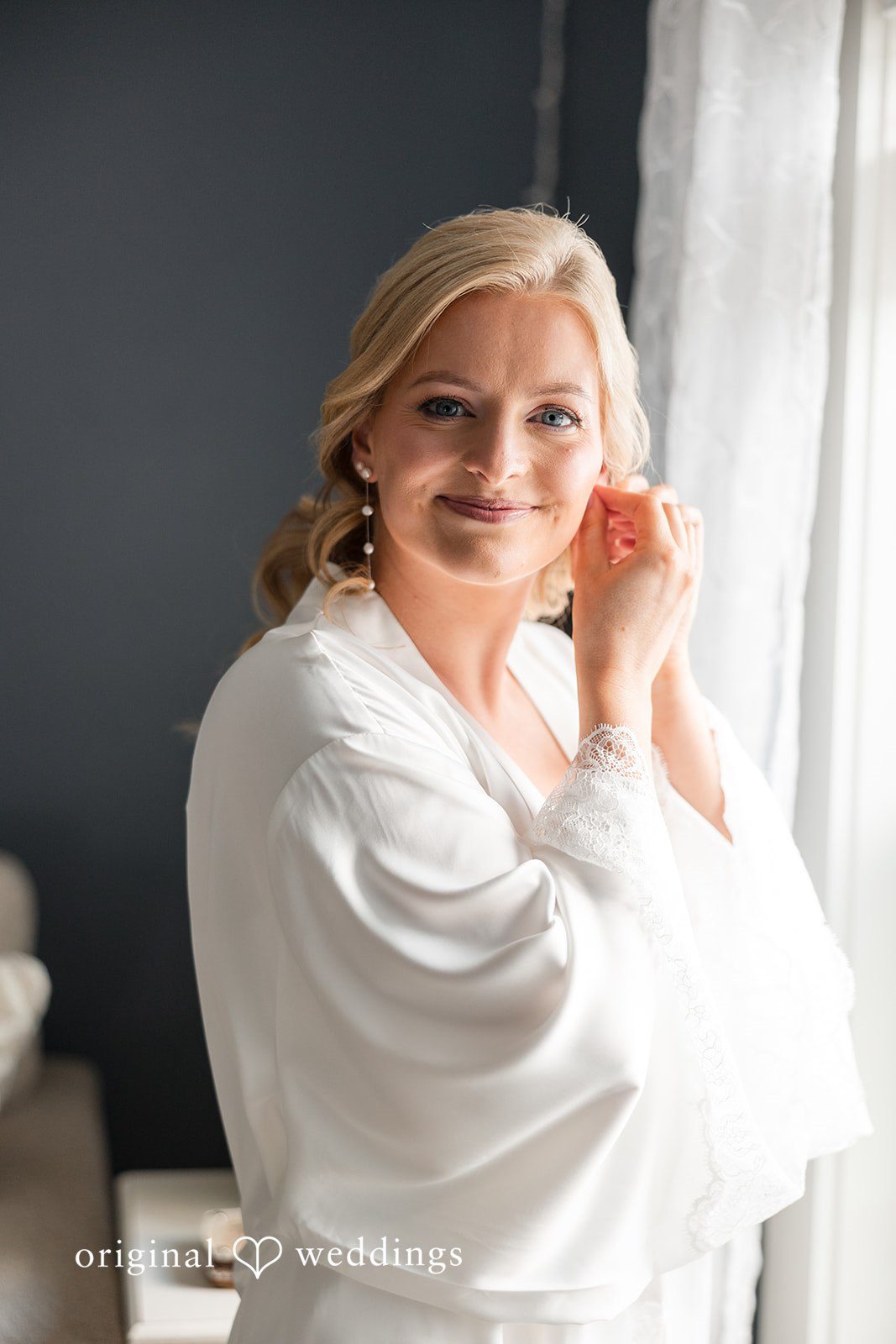 John + Emma Bride in a white robe putting on an earring while smiling.