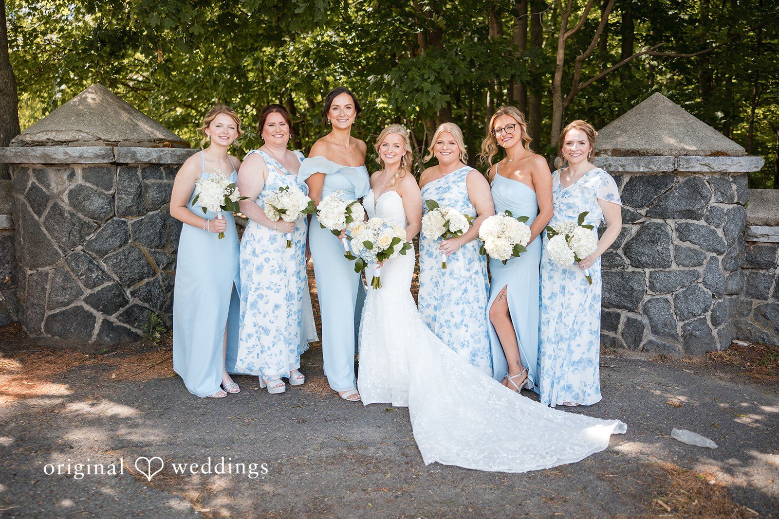John + Emma Bride and her bridesmaids posing in a line by a stone wall.