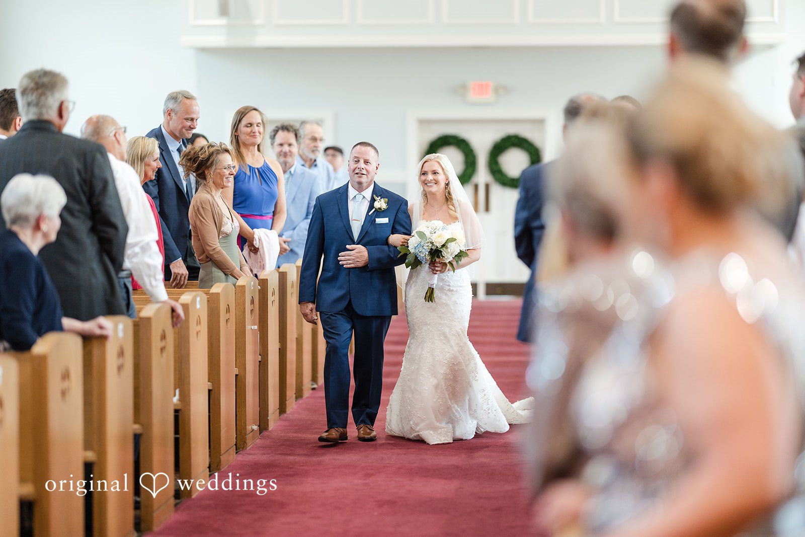 John + Emma Bride walking down the red carpet aisle of a church.