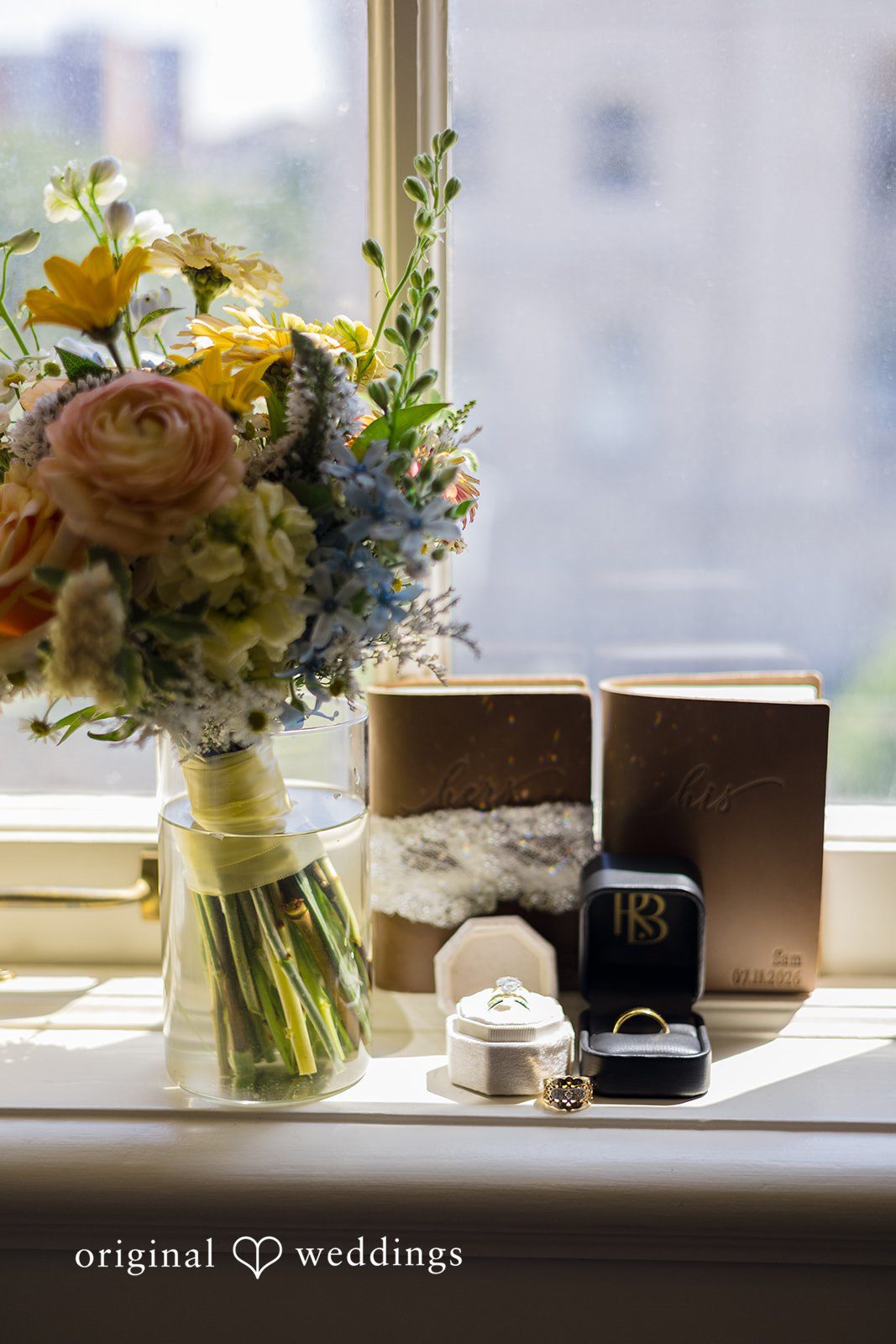 Wedding bouquet, vow books, and wedding rings displayed by the window at Memorial Union captured by Madison Wedding Photographers from Original Weddings.