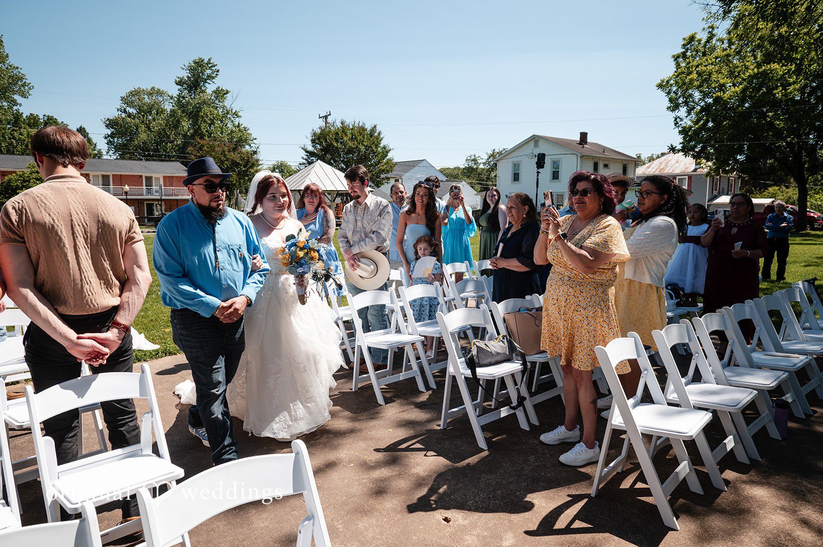 Joshua + Angela Wedding guests standing among rows of white chairs as a bride walks down the aisle.