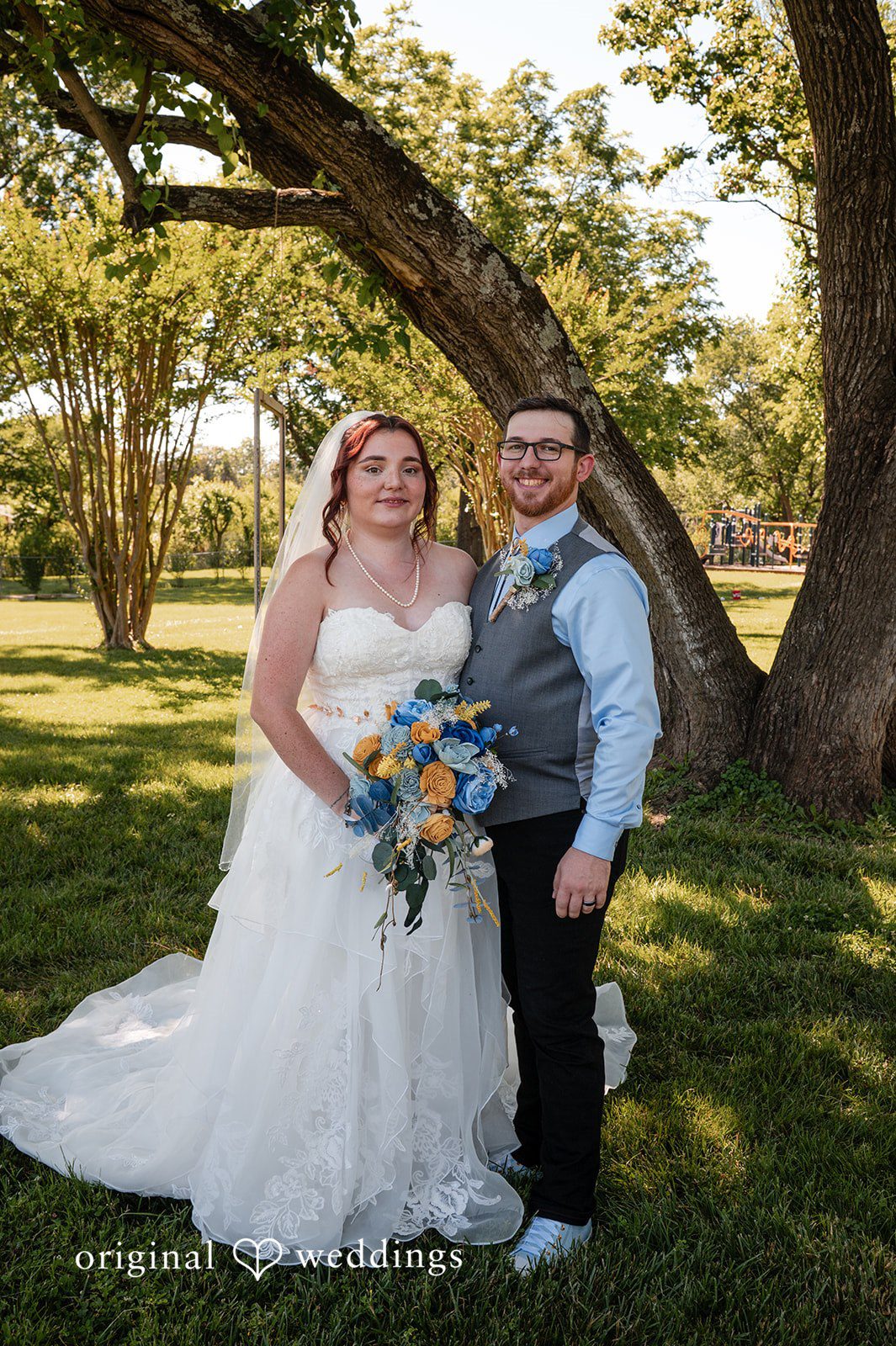 Joshua + Angela A bride and groom smile together while posing on a sunny green lawn.