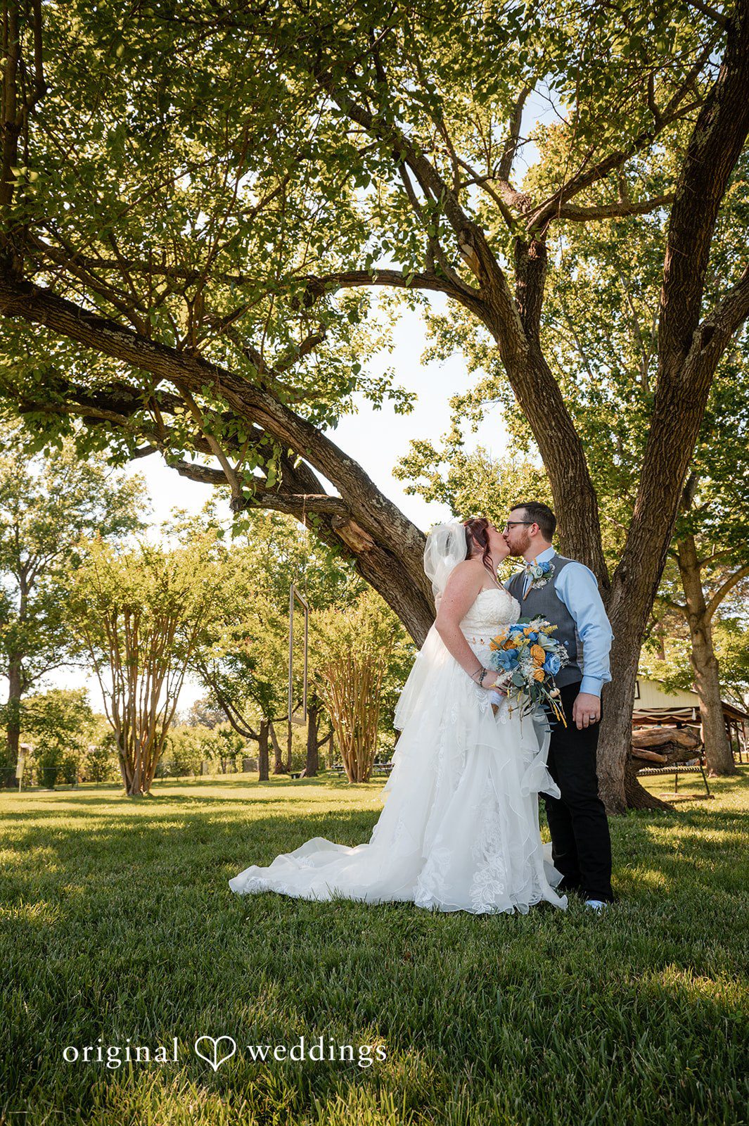 Joshua + Angela A bride and groom embrace on a grassy lawn shaded by large trees.
