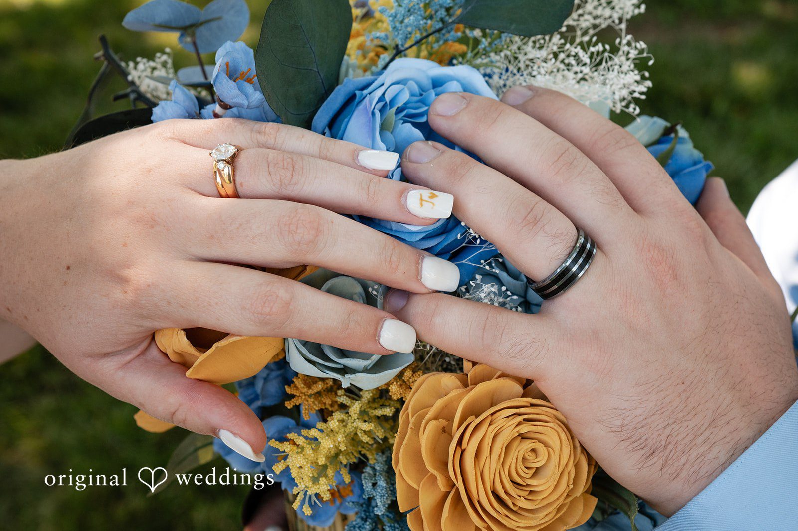 Joshua + Angela A close-up view of a bride and groom holding hands, showcasing their wedding rings and a colorful wooden floral bouquet.