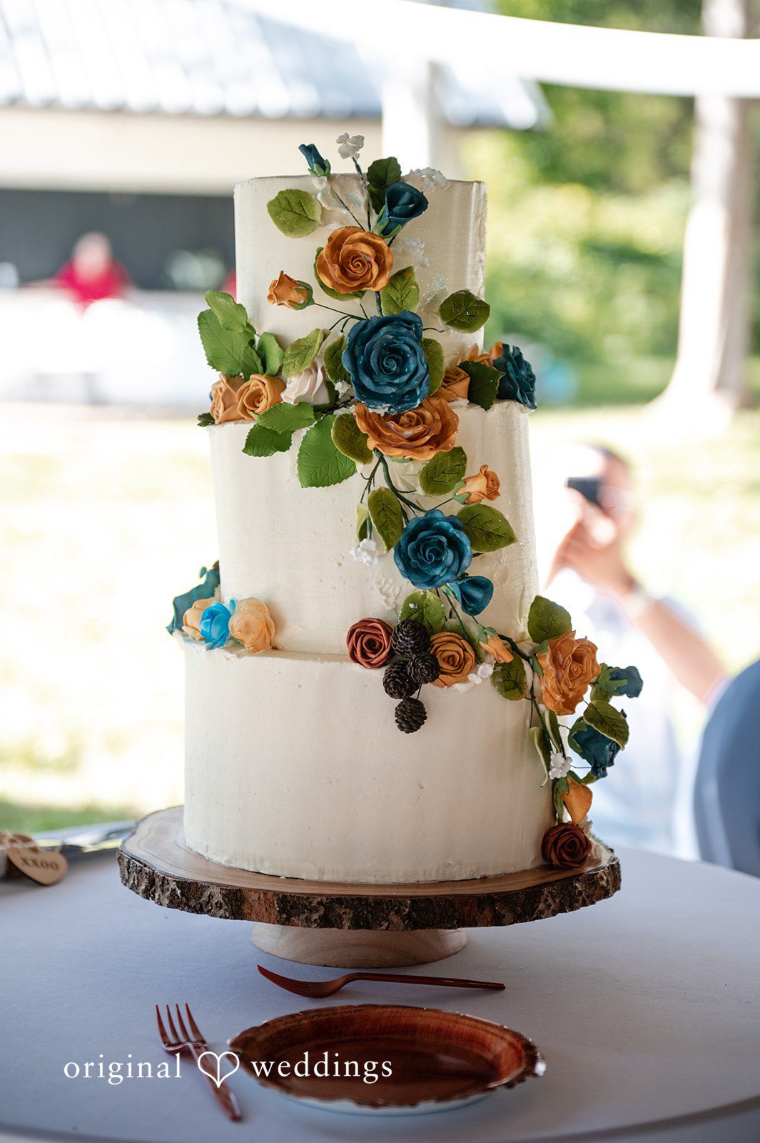 Joshua + Angela A three-tiered white frosted wedding cake decorated with blue, orange, and yellow flowers on a stand.