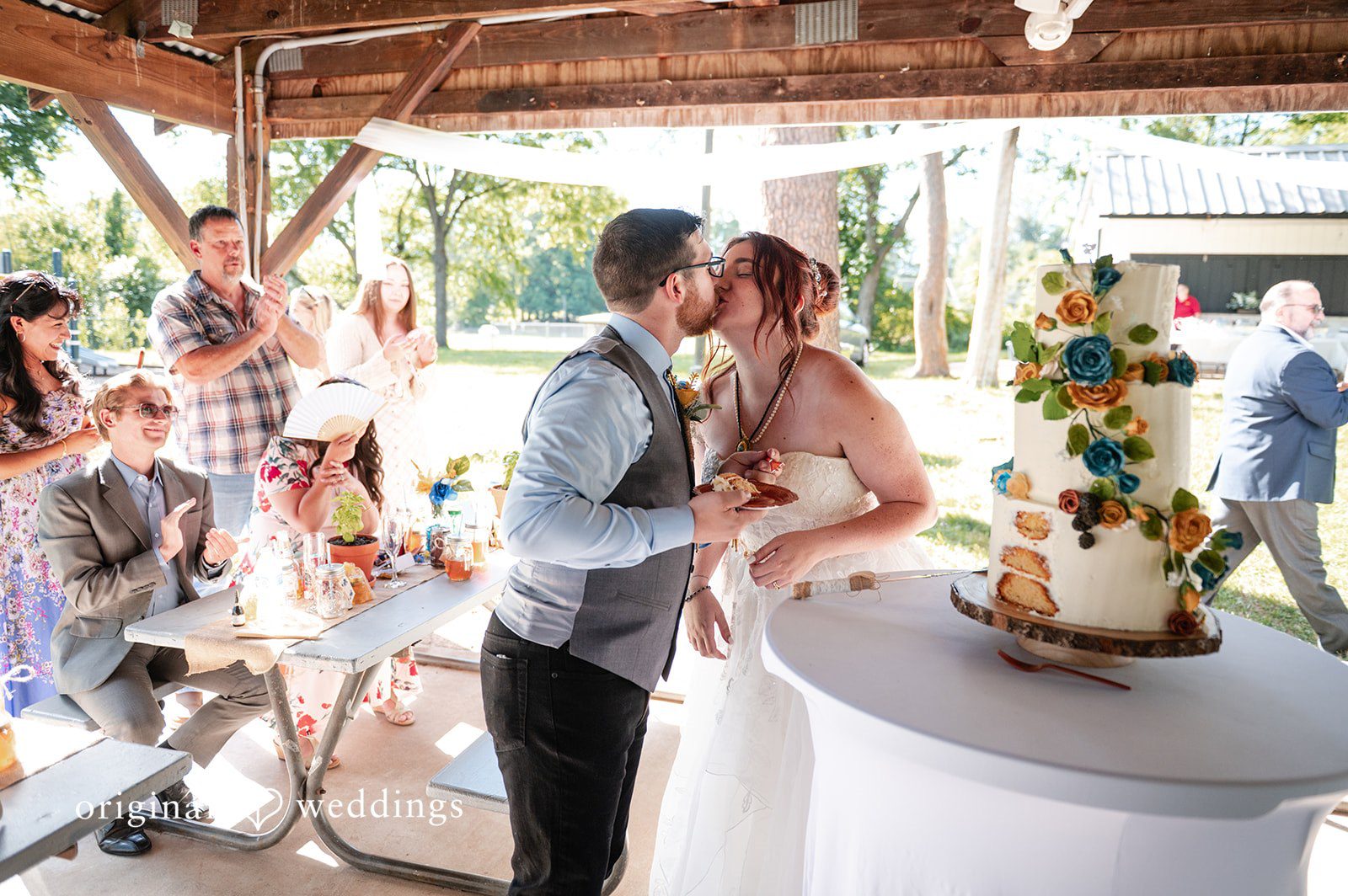 Joshua + Angela A bride and groom sharing a kiss next to their wedding cake table while guests watch.