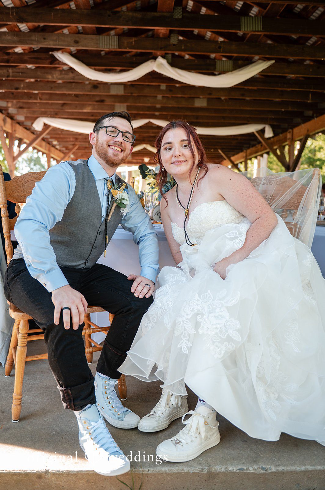 Joshua + Angela A bride and groom sitting together on chairs under a rustic pavilion, smiling at the camera.