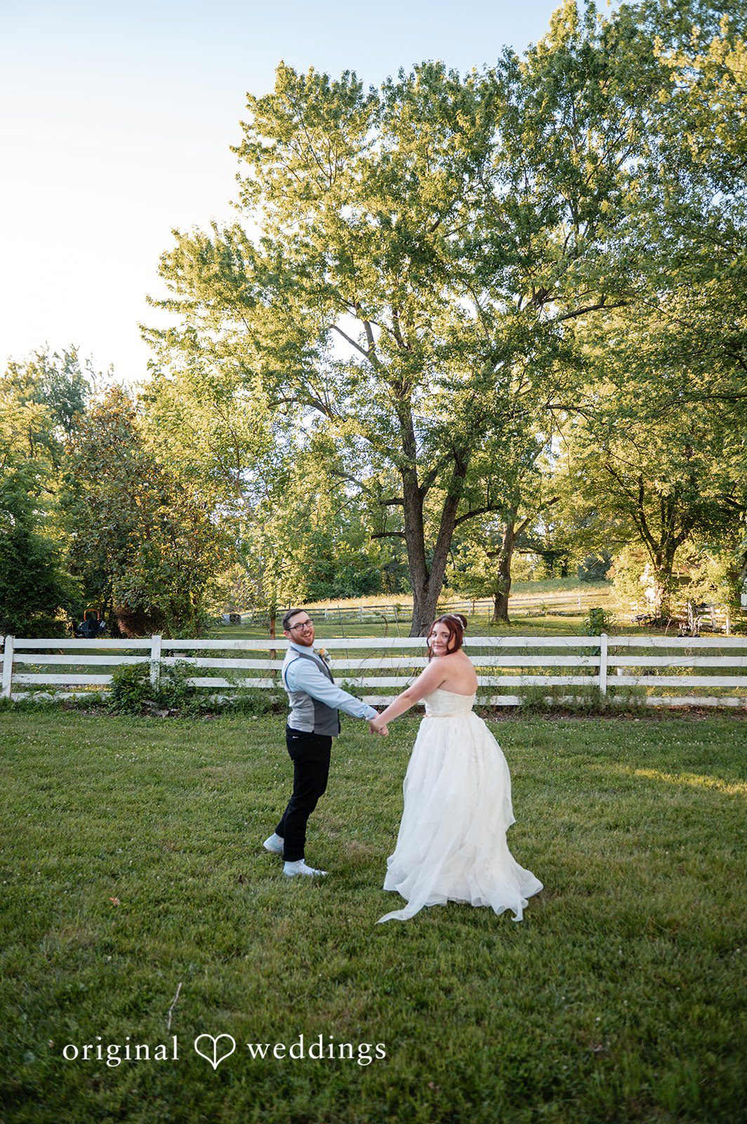 Joshua + Angela A bride and groom holding hands and dancing on a wide green lawn surrounded by trees.