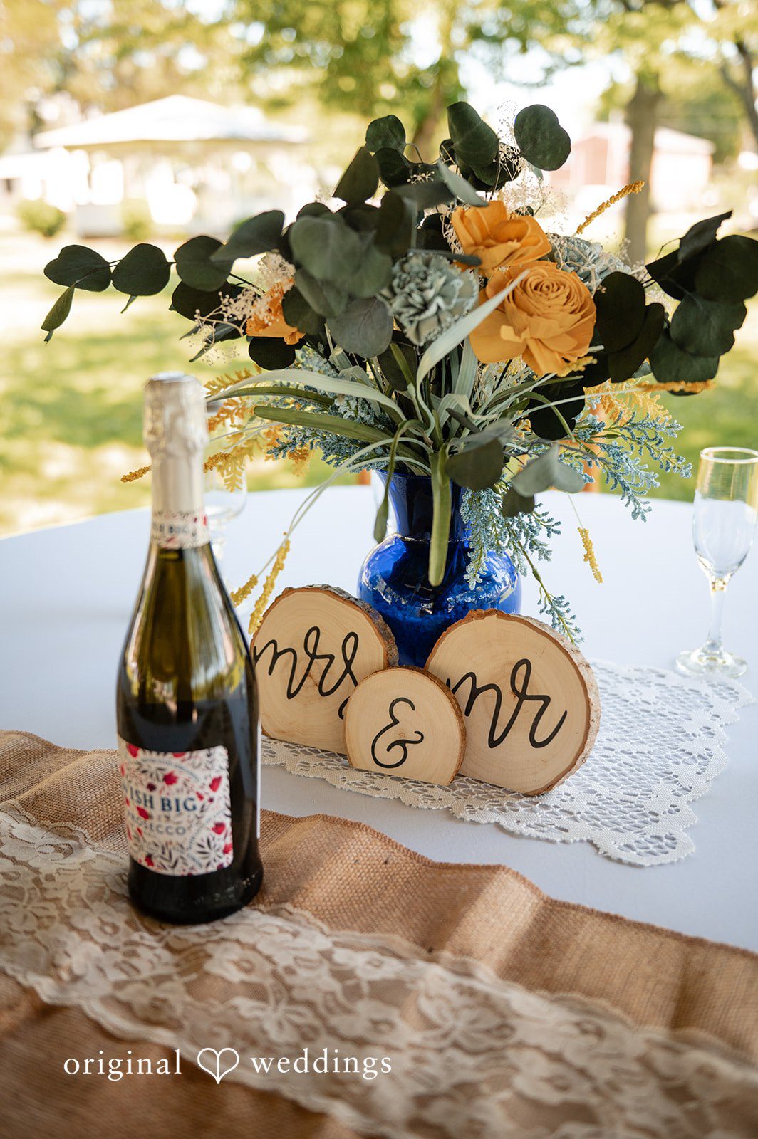 Joshua + Angela A rustic wedding reception table centerpiece featuring a bottle of champagne, floral arrangements, and wooden "Mr. & Mrs." signs.