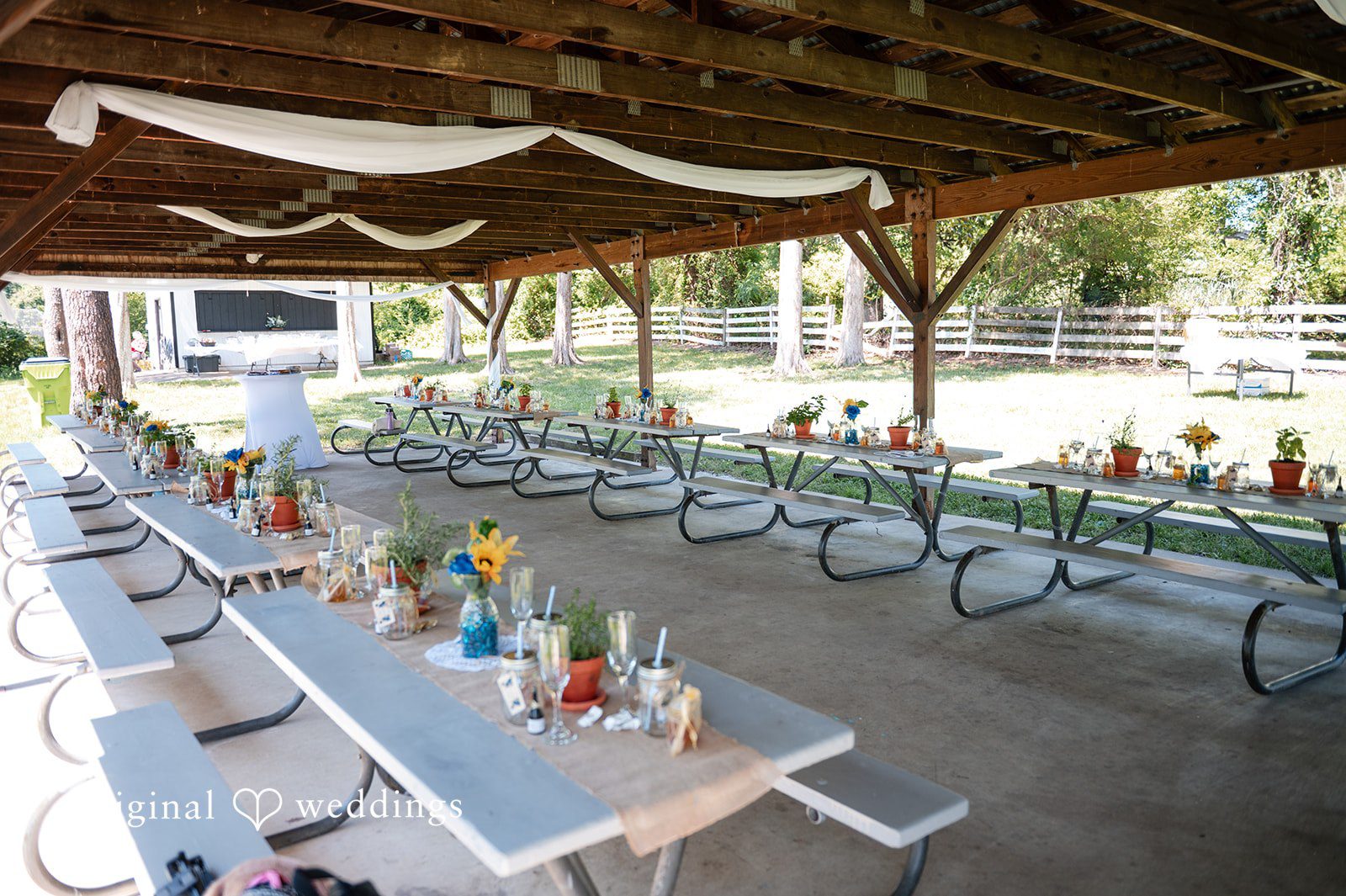 Joshua + Angela Long wooden picnic tables arranged for a wedding reception under a covered patio structure.