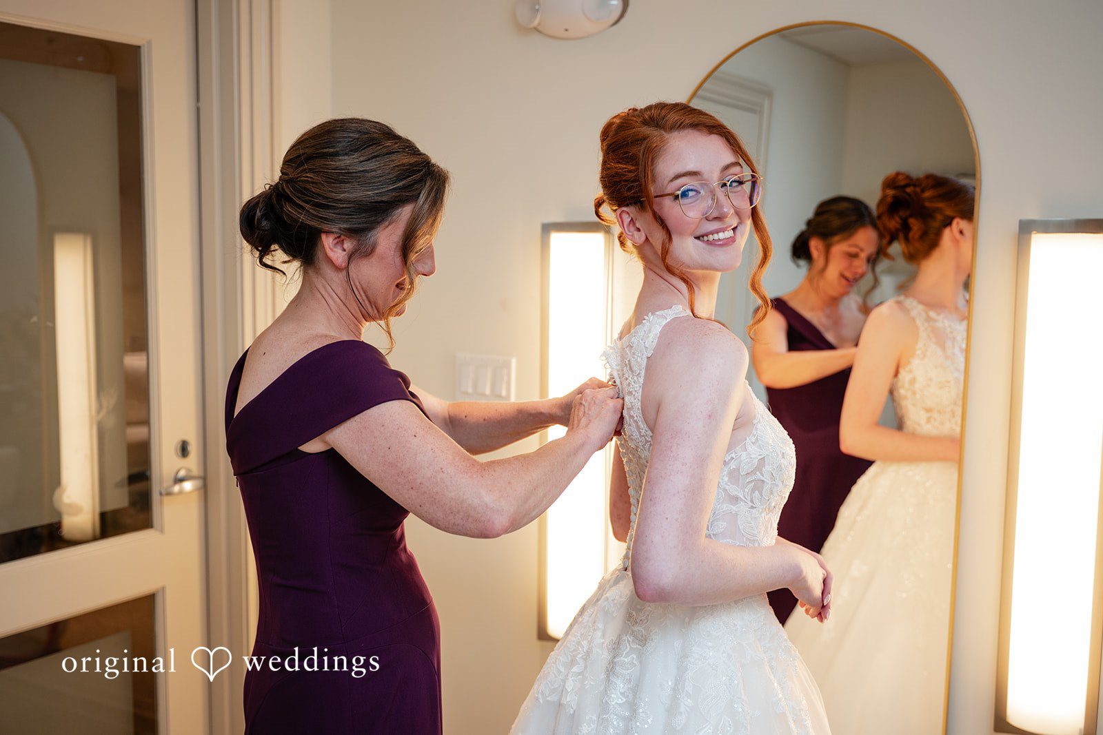 Aislyn + Alex Woman in a purple dress helping a redhead bride zip up her white wedding gown.