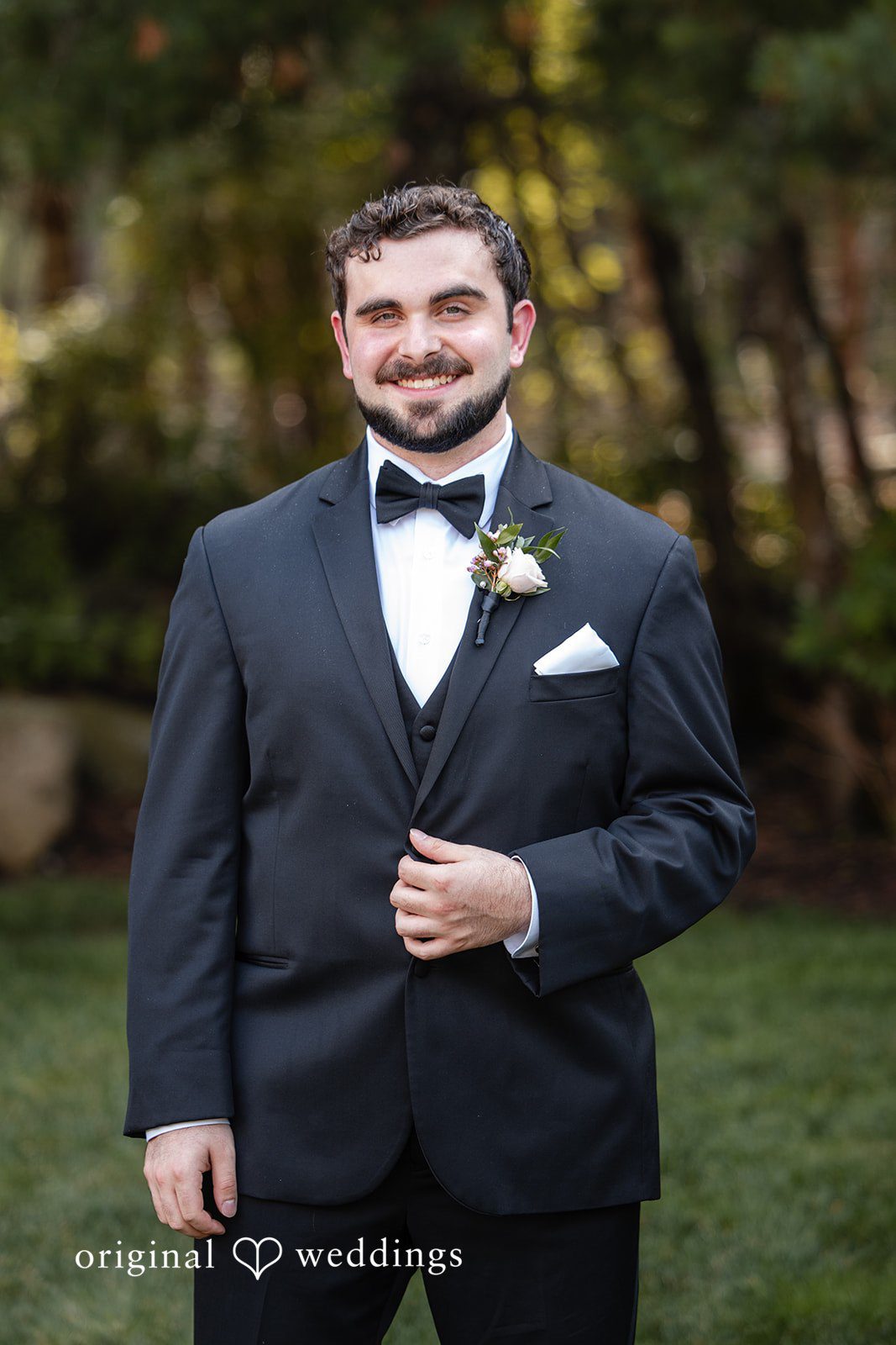 Aislyn + Alex Portrait of a groom smiling in a black tuxedo and bow tie against a blurred background.