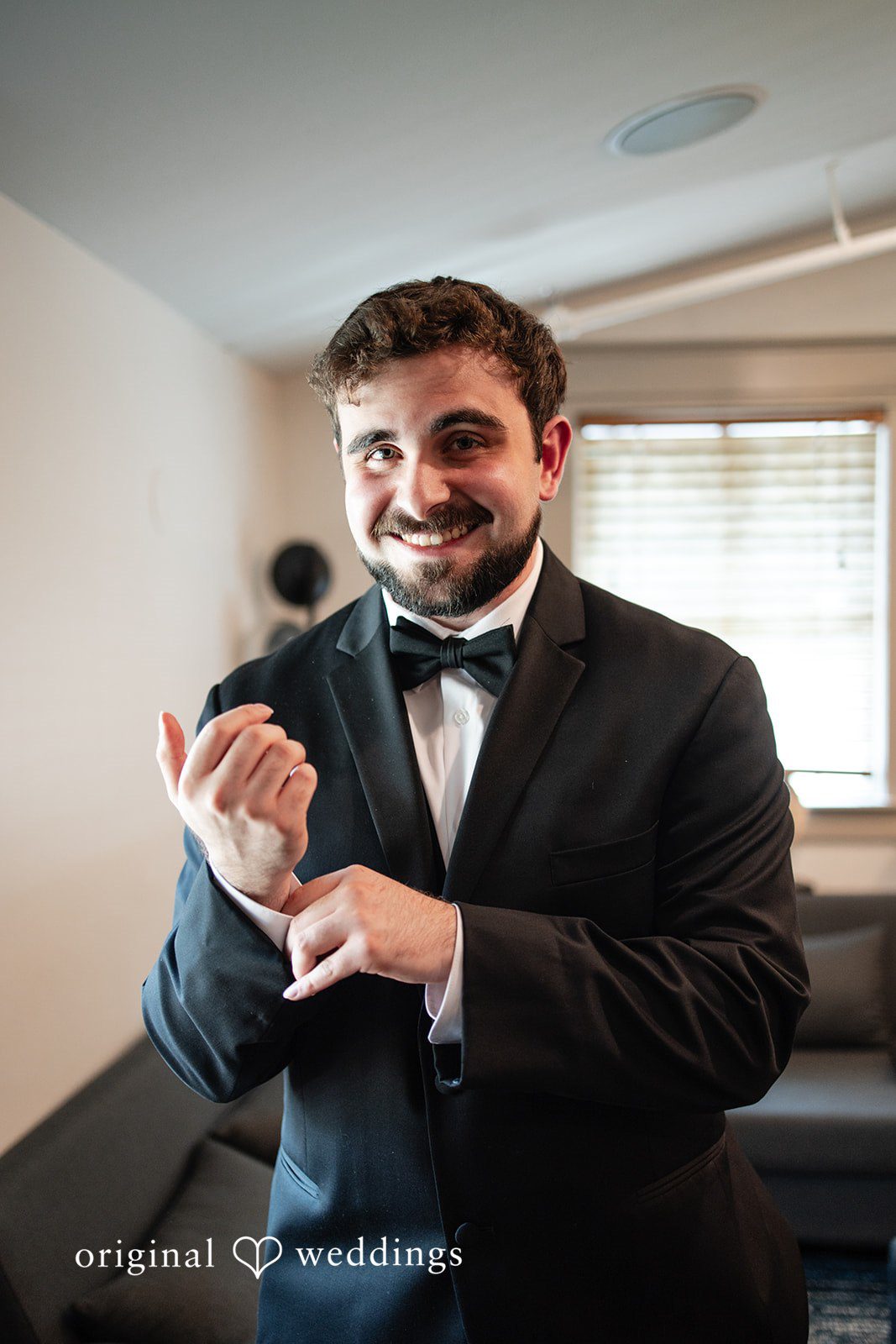 Aislyn + Alex Groom in a black tuxedo and bow tie smiling while adjusting his cuffs.