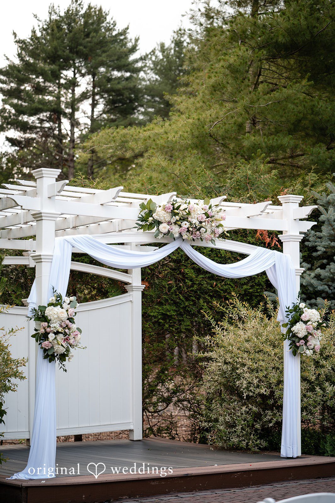 Aislyn + Alex White wedding arch decorated with fabric and flowers on a lawn.