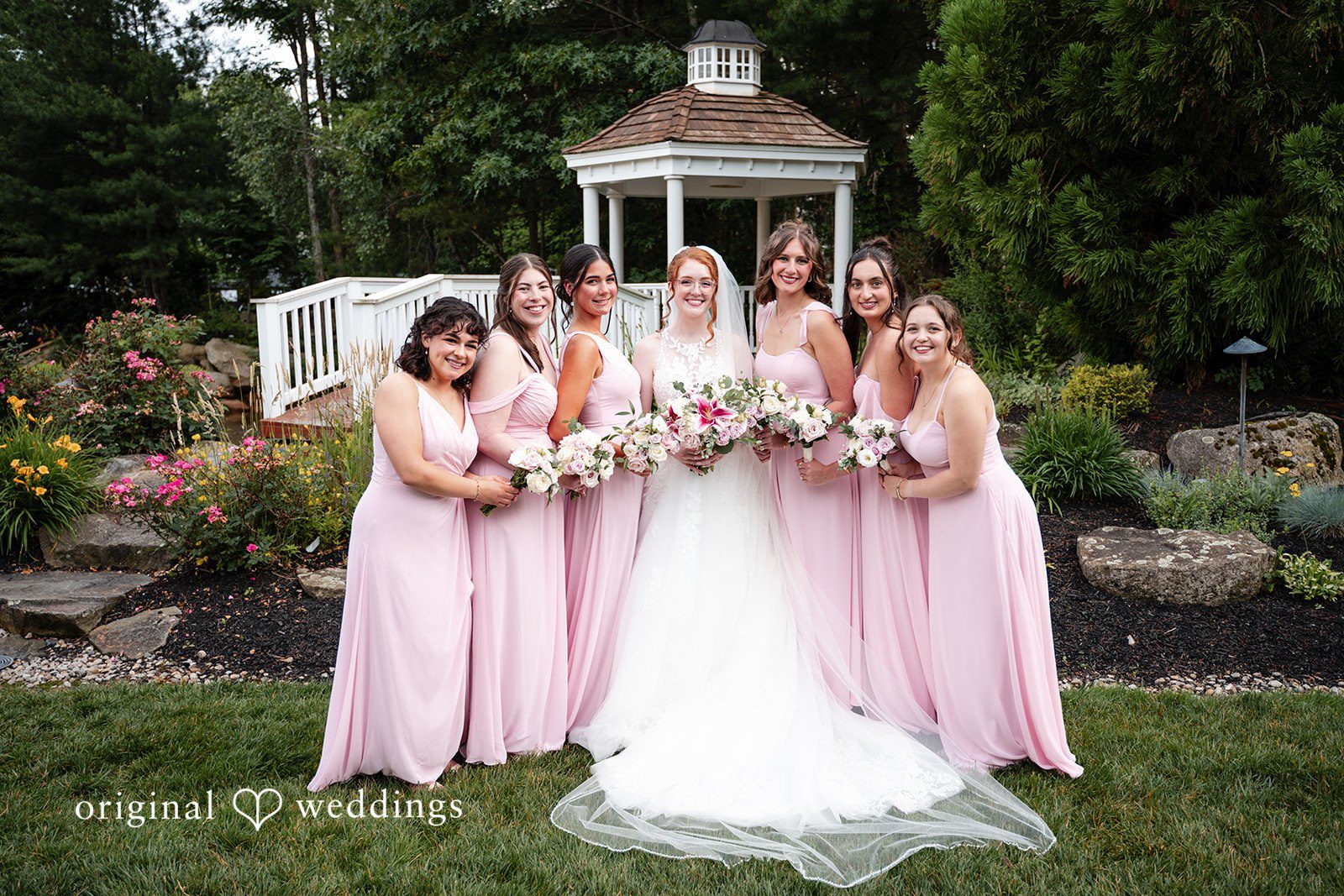 Aislyn + Alex Bride posing with her bridesmaids in pink dresses on a lawn near a gazebo.