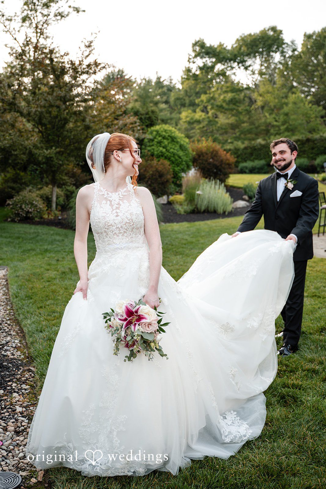 Aislyn + Alex Groom holding the train of the bride's white gown as they walk through a garden.