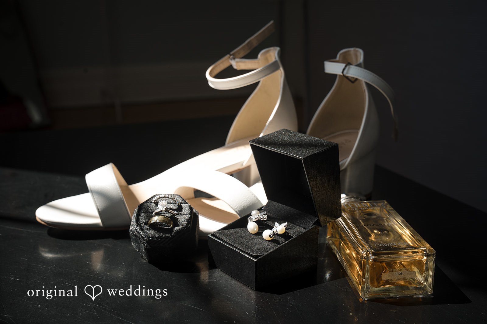 Ashley + Jonathan Bridal high-heeled shoes arranged on a dark surface next to a perfume bottle.