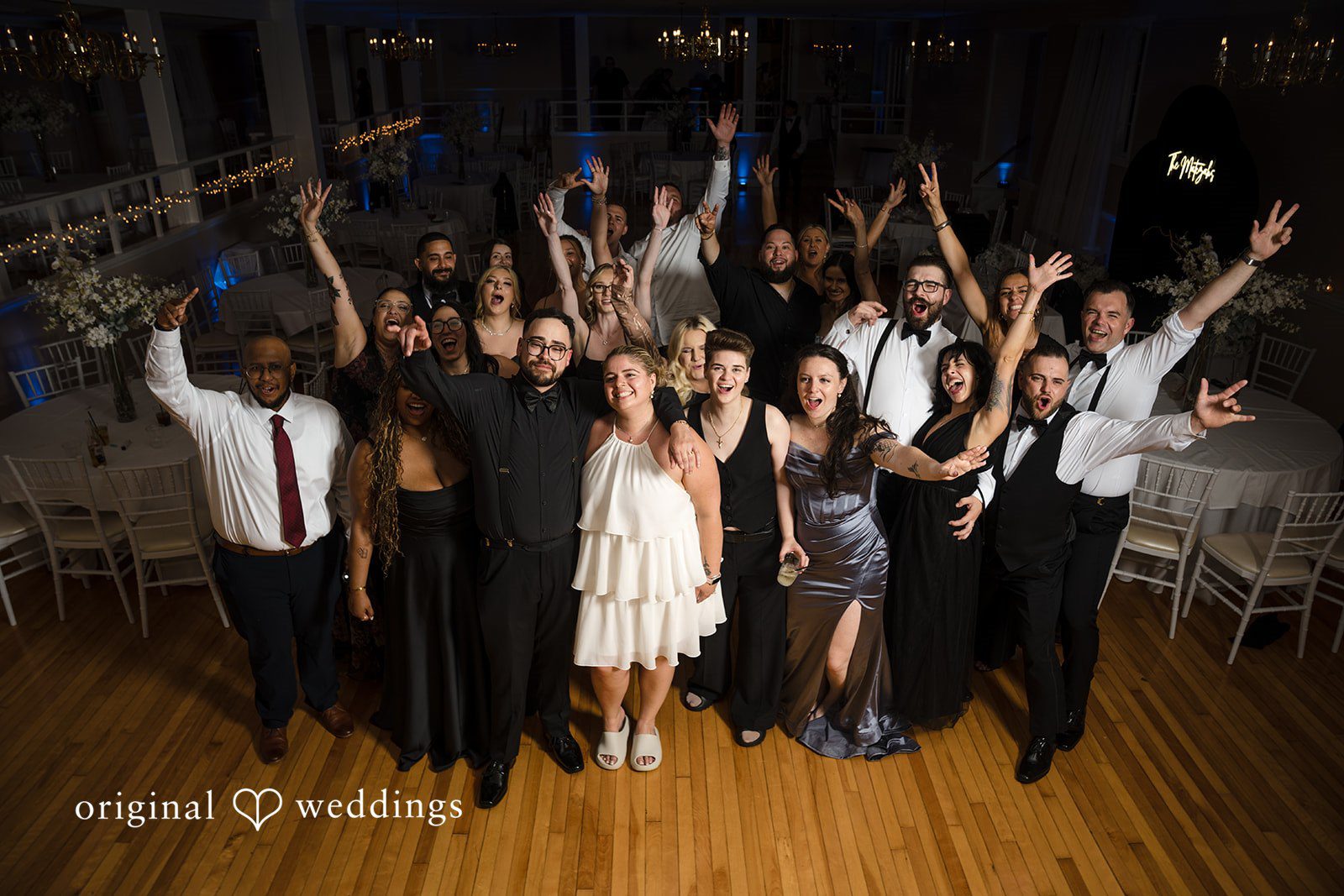 Ashley + Jonathan A large group of wedding guests cheering and raising their hands together on the dance floor.