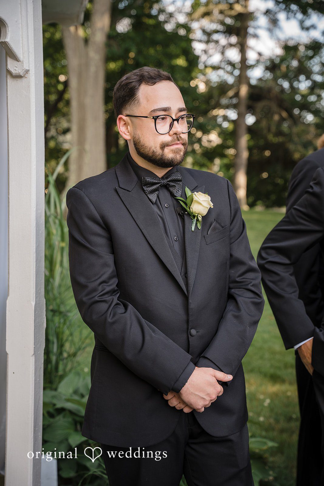 Ashley + Jonathan Groom posing in his black tuxedo on a lawn.