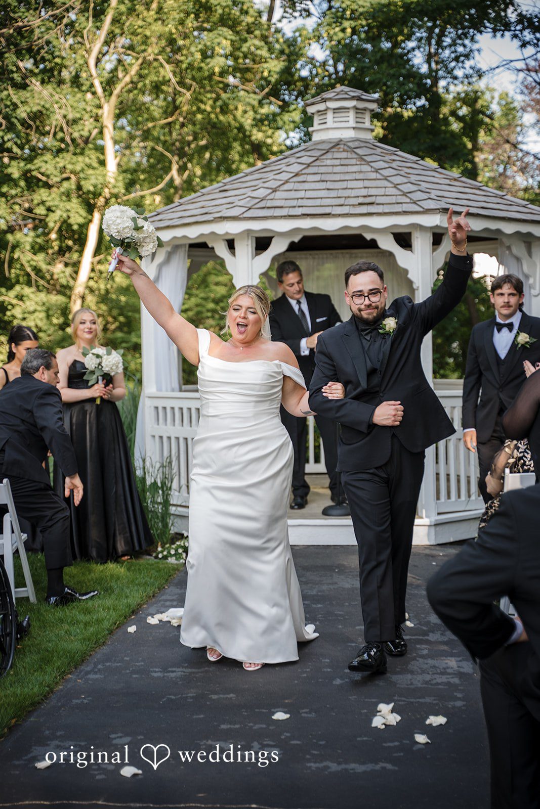 Ashley + Jonathan A newlywed bride and groom celebrating with raised arms while walking down an aisle.