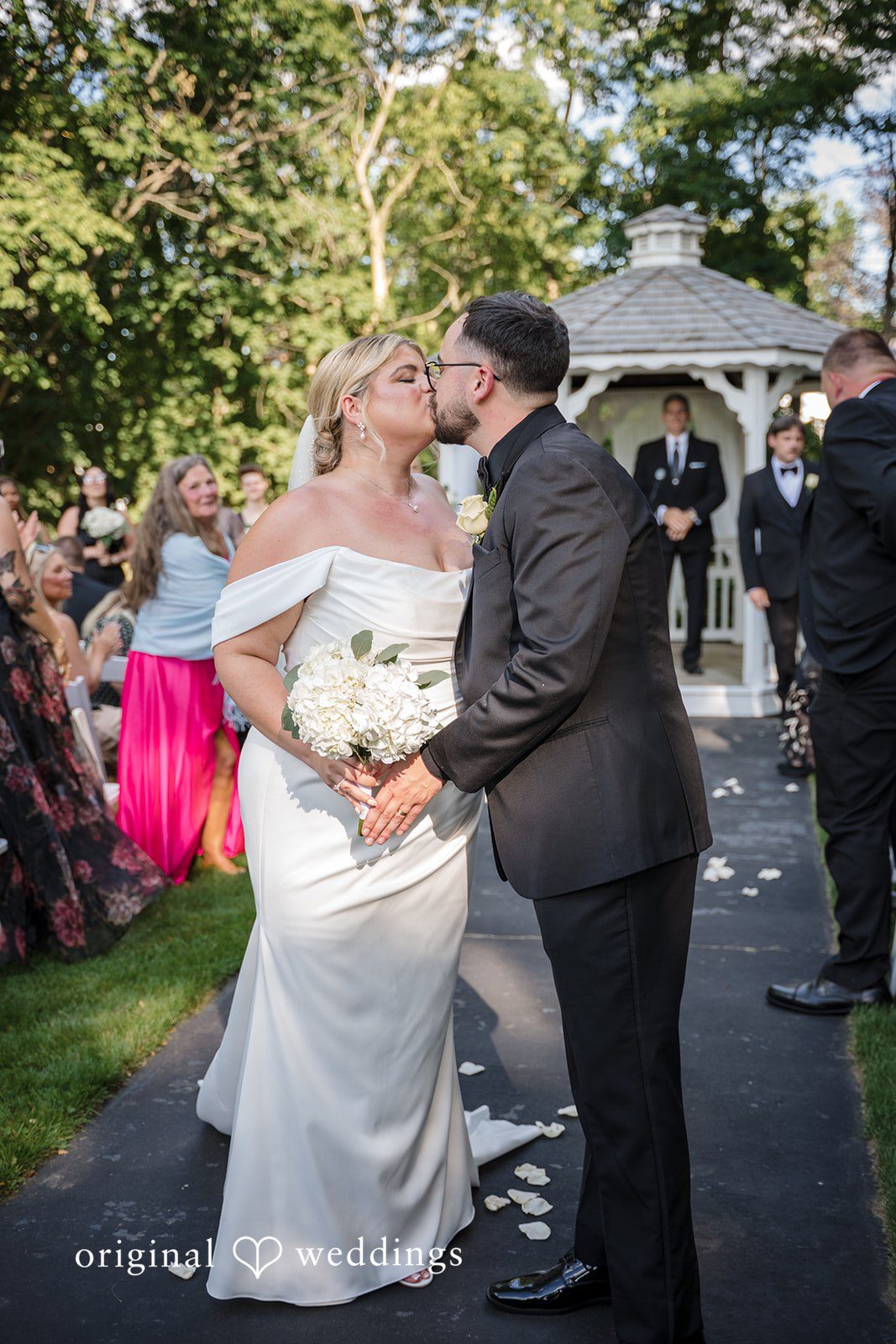 Ashley + Jonathan A bride and groom kissing on a walkway surrounded by wedding guests.