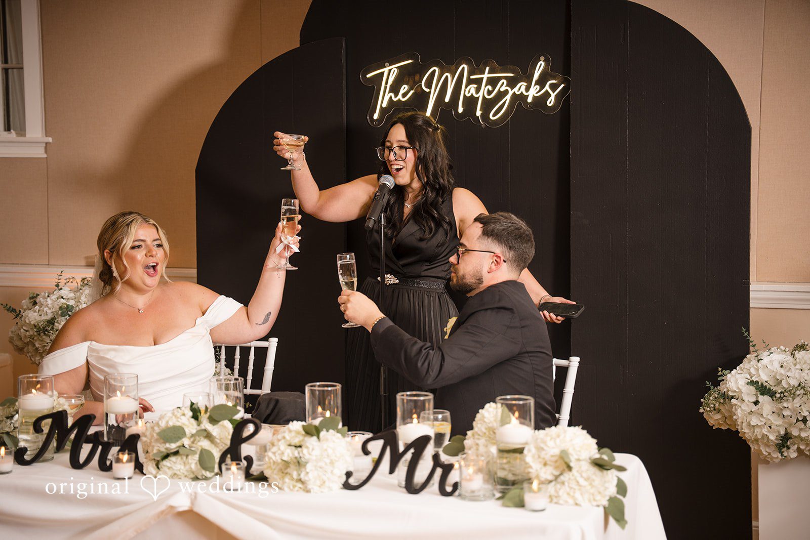 Ashley + Jonathan A bride sitting at her wedding reception head table while bridesmaids celebrate behind her.