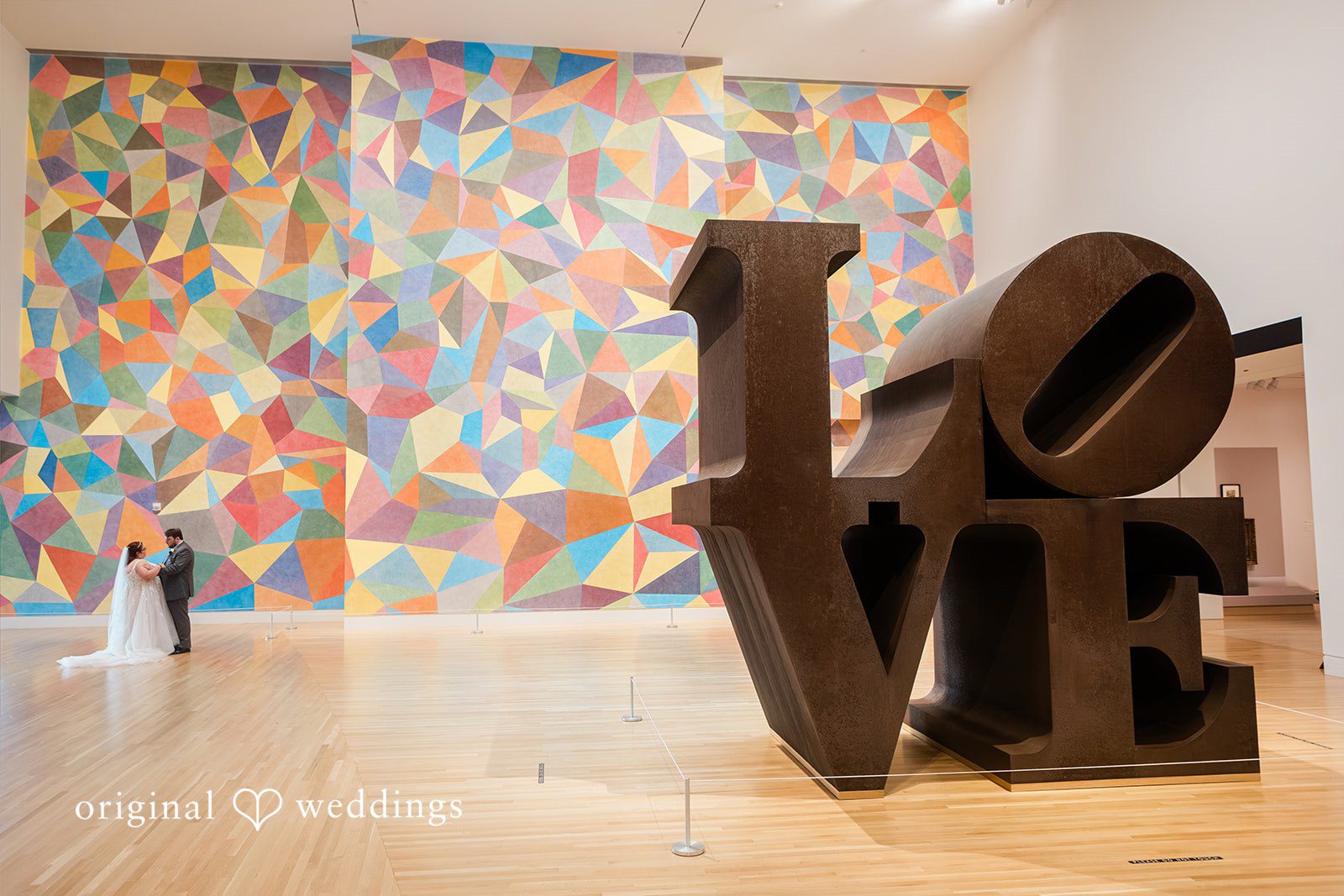 Grace + Nicholas A bride and groom standing beside a large, iconic LOVE sculpture in front of a colorful geometric wall.