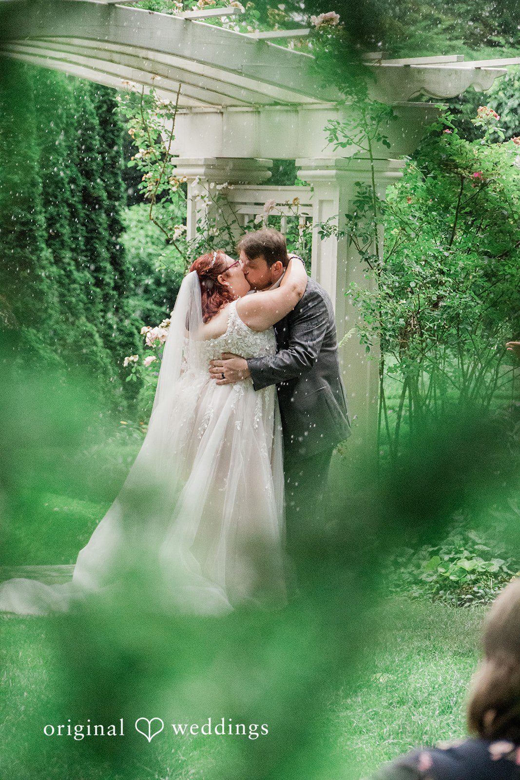 Grace + Nicholas A bride and groom sharing a romantic kiss framed by green foliage.
