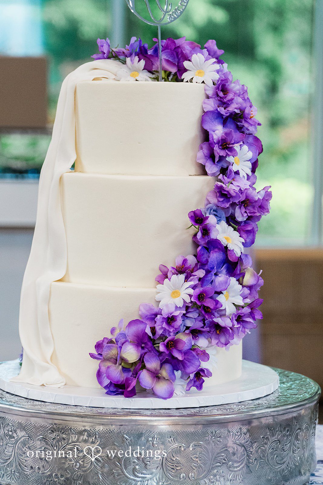 Grace + Nicholas A close-up of a white three-tiered wedding cake decorated with a cascading drape of purple flowers.