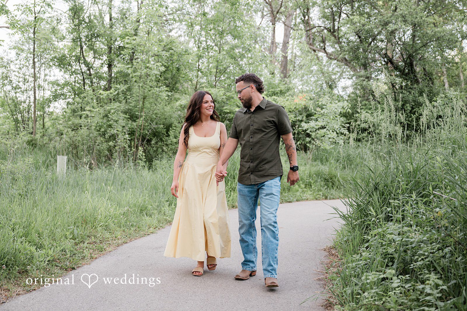 Andrea + Jordan A couple walking hand-in-hand down a paved path surrounded by lush trees and greenery.