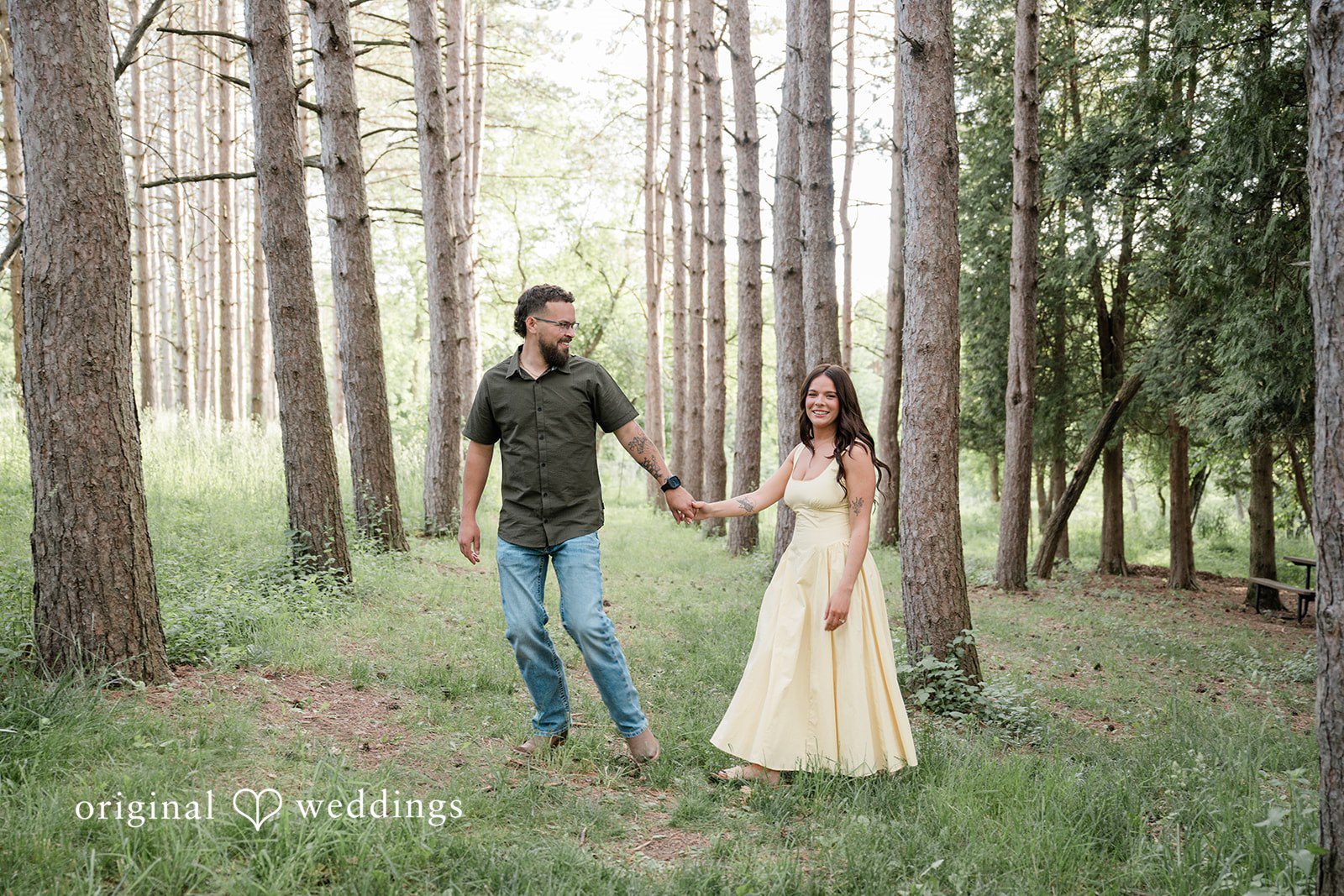 Andrea + Jordan A couple holding hands and walking together through a grove of tall pine trees.