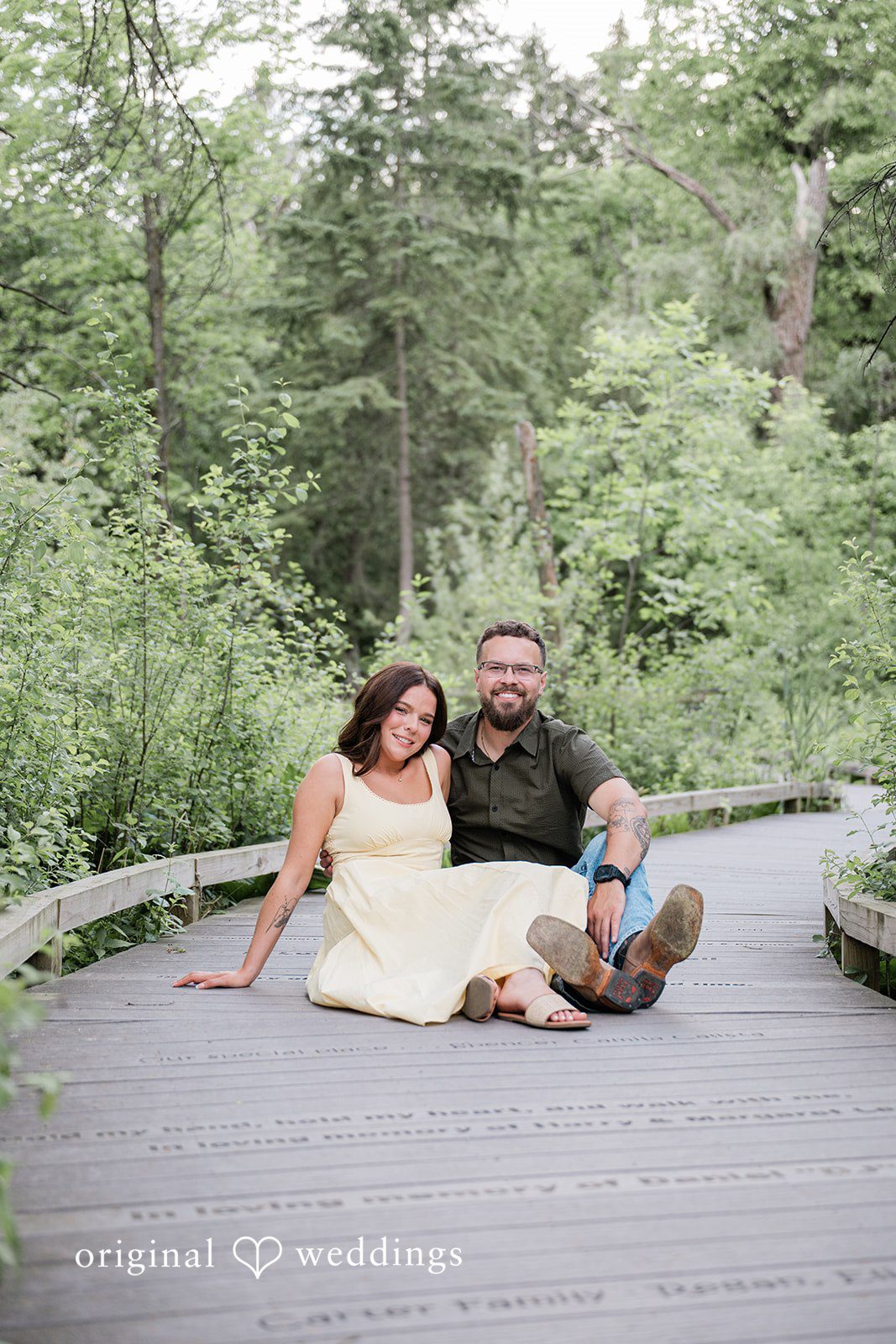 Andrea + Jordan A couple relaxing together on a wooden boardwalk path winding through a wooded area.