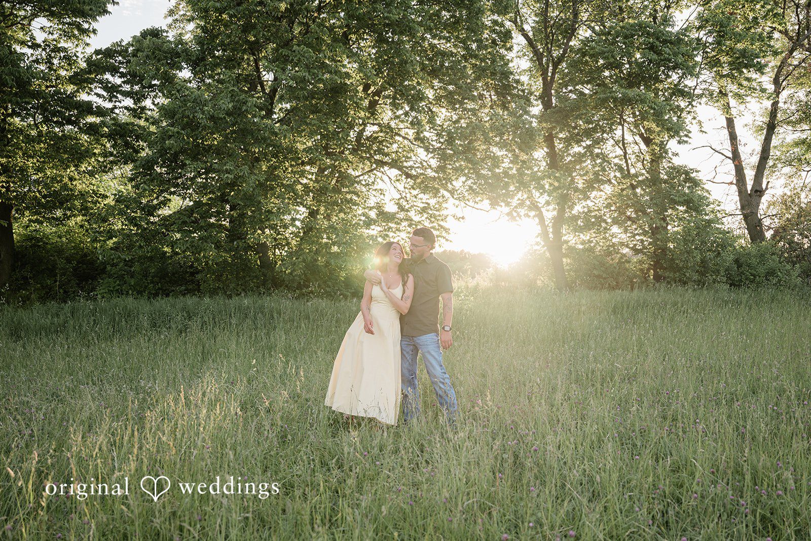 Andrea + Jordan A couple sharing a close embrace in a grassy field with bright sunlight filtering through the trees behind them.