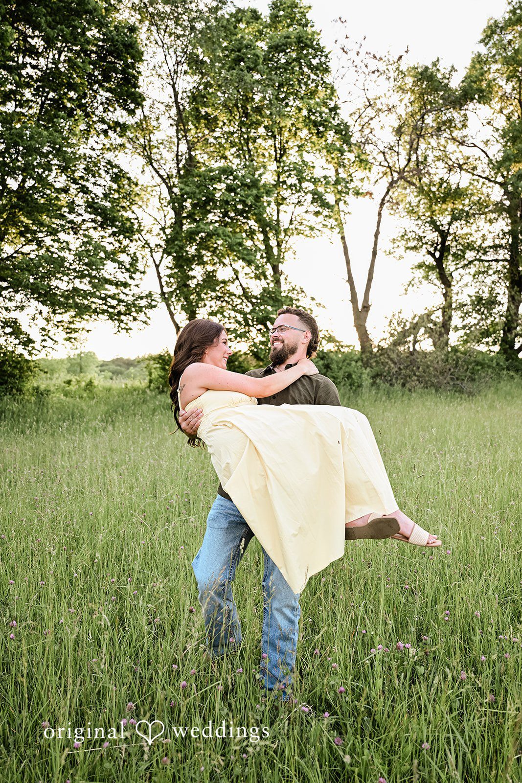 Andrea + Jordan A man joyfully lifting and carrying a woman across a grassy meadow.