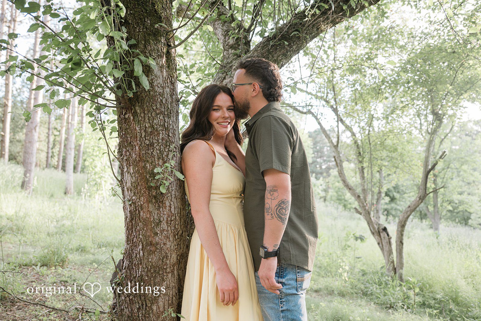 Andrea + Jordan A couple standing close together and smiling under the shade of tall forest trees.