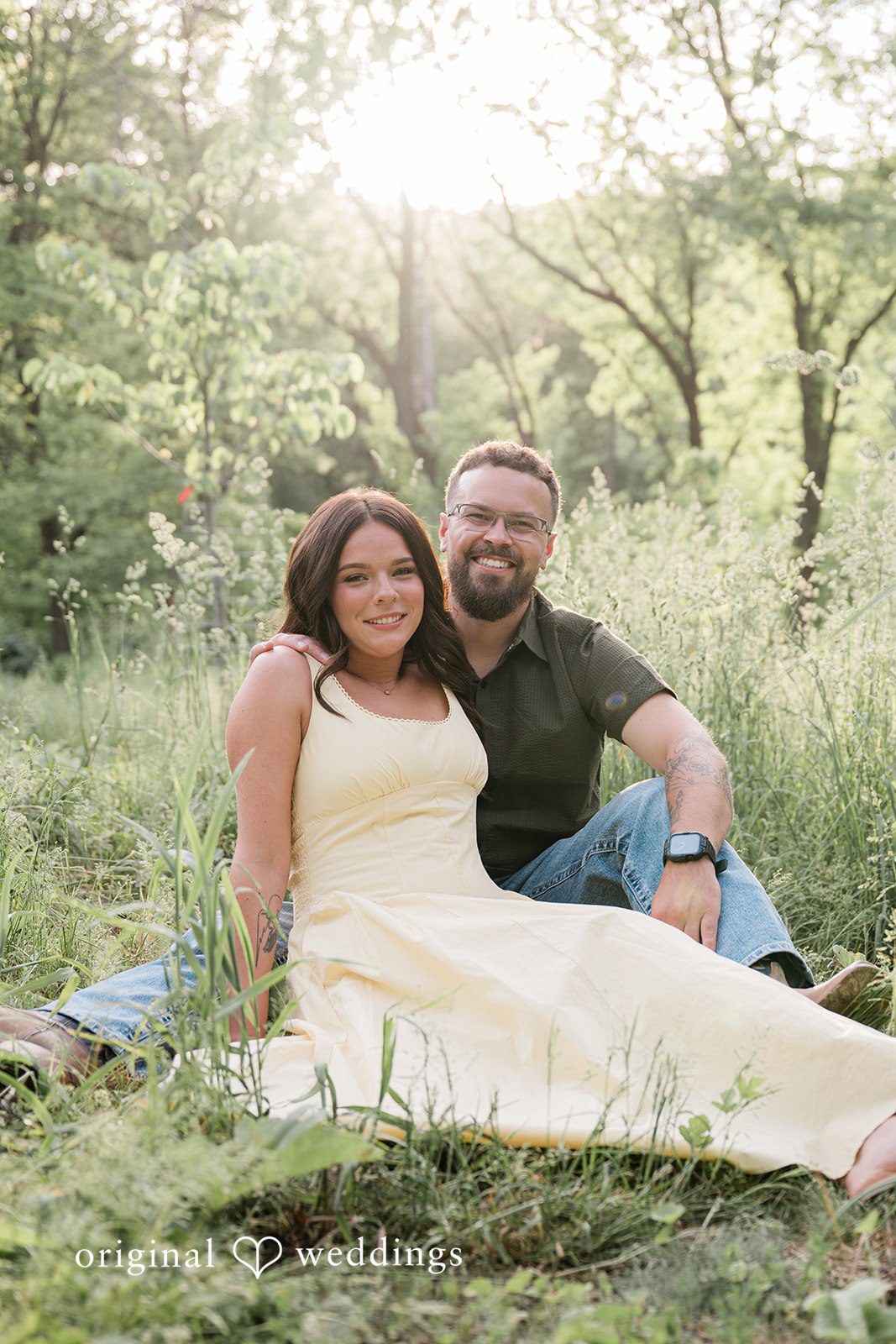 Andrea + Jordan A couple sitting together on a grassy slope with trees in the background.
